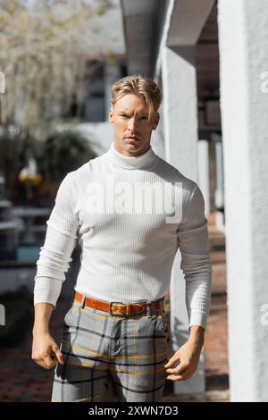 Un bel homme blond pose dans un col roulé blanc et un pantalon à carreaux sur une rue ensoleillée de Floride. Banque D'Images