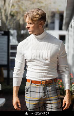 Un bel homme blond pose dans un col roulé blanc et un pantalon à carreaux dans une rue de Floride. Banque D'Images