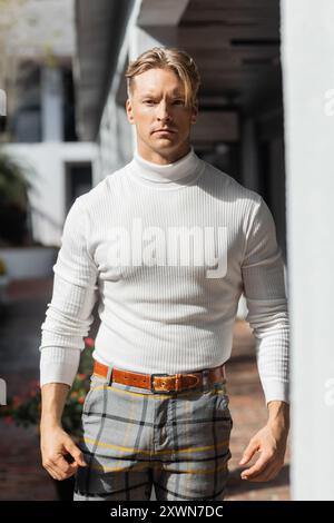 Un bel homme blond pose dans un pull à col roulé blanc et un pantalon à carreaux sur une rue ensoleillée de Floride. Banque D'Images