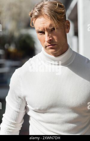 Un bel homme blond dans un col roulé blanc pose sur une rue ensoleillée de Floride. Banque D'Images