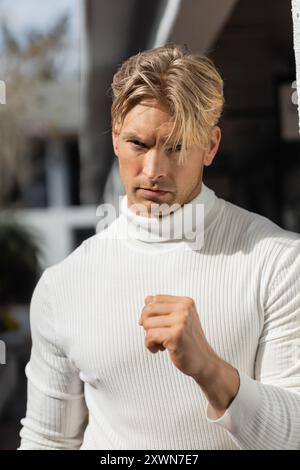 Un bel homme blond avec une expression déterminée pose pour une photo tout en portant un pull blanc à col roulé dans une rue de Floride. Banque D'Images