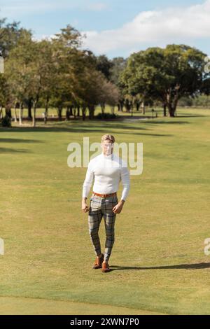 Un homme blond à col roulé blanc et pantalon à carreaux marche à travers un parc verdoyant à Miami. Banque D'Images