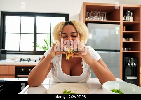 Jeune femme afro-américaine courbée dans la cuisine appréciant manger gros hamburger Banque D'Images