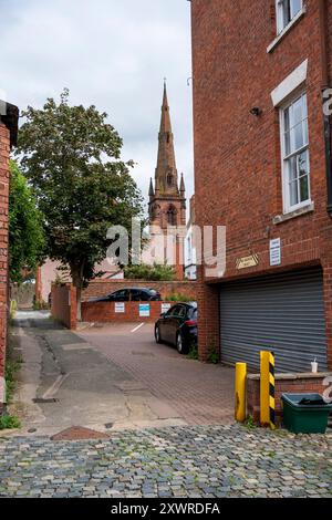 Ruelle étroite menant à une église avec une grande flèche, flanquée de bâtiments en briques par temps nuageux. Banque D'Images