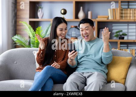 Famille asiatique appréciant les nouvelles passionnantes sur un téléphone tout en étant assis sur un canapé à la maison. Couple d'âge mûr exprimant le bonheur et la surprise, mettant en valeur la technologie et la communication dans un cadre confortable. Banque D'Images