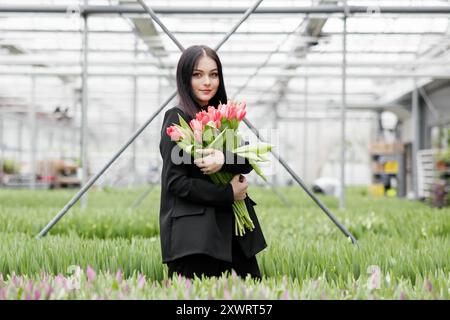 Jeune femme debout dans une grande serre et tenant des tulipes en fleurs dans ses mains. Banque D'Images