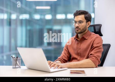 Jeune homme professionnel avec des lunettes profondément concentré sur le travail d'ordinateur portable dans le bureau moderne. Démontrer la concentration et la détermination. Le téléphone à proximité suggère le multitâche. Atmosphère de productivité Banque D'Images