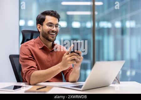 Un professionnel souriant assis au bureau utilise un smartphone tandis que son ordinateur portable et ses ordinateurs portables sont à proximité. Porter des lunettes et une tenue décontractée, montre une expression positive, illustrant la connectivité Banque D'Images