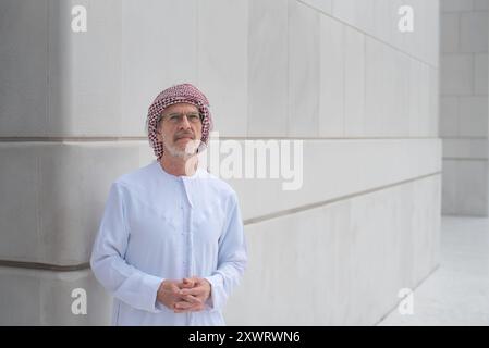 Homme arabe âgé se tient confiant portant des vêtements traditionnels kandora et foulard, posant contre un bâtiment moderne et minimaliste avec blanc, propre l Banque D'Images