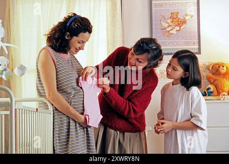 Une femme enceinte admire les vêtements de bébé tout en étant assistée par sa mère et surveillée par sa fille dans une crèche chaleureusement décorée. Banque D'Images