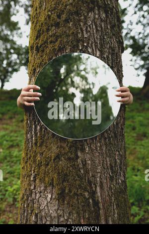 Concept de nature et d'environnement. Mains de femme tenant un miroir rond contre un arbre, reflétant la verdure. L'image contient du bruit et du flou de mouvement Banque D'Images