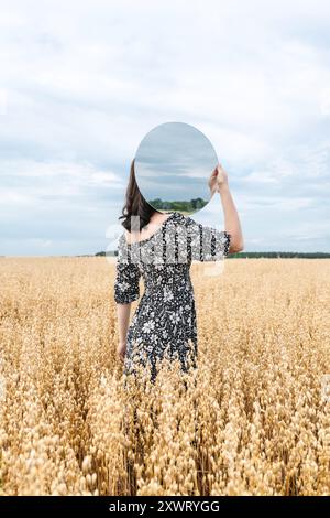 Une femme au feuillage robe noire et blanche tient un miroir rond reflétant le ciel dans un champ de blé doré. L'image contient du bruit et du flou de mouvement. Sele Banque D'Images