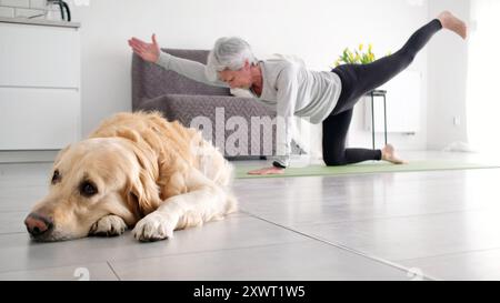 Femme âgée active faisant du yoga à la maison tandis qu'Un chien Golden Retriever repose sur le sol Banque D'Images