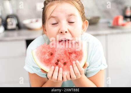 Petite adolescente mord pastèque avec ses yeux fermés avec plaisir. Délicieux dessert d'été mangé par une petite fille dans la cuisine. Haute qualité p Banque D'Images