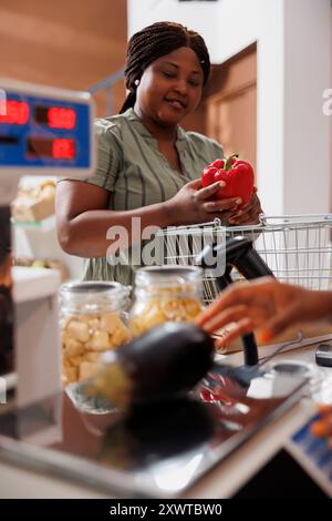 Happy Black Woman fait ses courses sur un marché alimentaire local pour des produits frais et biologiques. Elle sourit tandis que le commerçant l'aide à peser ses produits frais. Il y a une variété de légumes disponibles. Banque D'Images