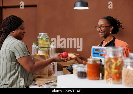 Femme afro-américaine commerçante aide un client à la caisse d'un marché alimentaire. Produits frais et produits biologiques disponibles à l'achat. Banque D'Images