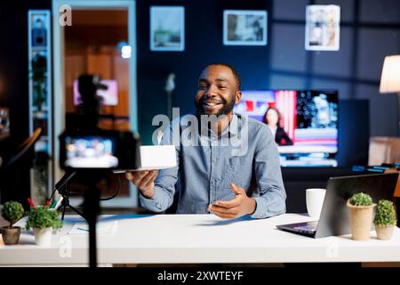 Créateur de contenu souriant à la maison avec un produit en attente d'être déballé devant lui pendant qu'il fait l'introduction vidéo. Influenceur tendance réalisant le déballage des séquences pour les téléspectateurs Banque D'Images