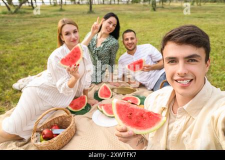 Jeune homme avec pastèque et ses amis prenant selfie sur pique-nique dans le parc, gros plan Banque D'Images