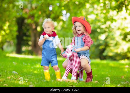 Petit garçon et fille habillés comme cow-boy et cow-girl jouant avec jouet cheval à bascule dans le parc. Les enfants jouent à l'extérieur. Enfants en costumes d'Halloween Banque D'Images