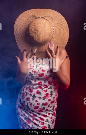 Belle femme debout portant une robe colorée avec un chapeau couvrant toute sa tête. Isolé sur fond fumé coloré. Banque D'Images