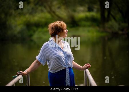 Une femme se tient sur un quai, regardant attentivement les eaux calmes d'un lac, encadrées de verdure sous un soleil éclatant. Banque D'Images