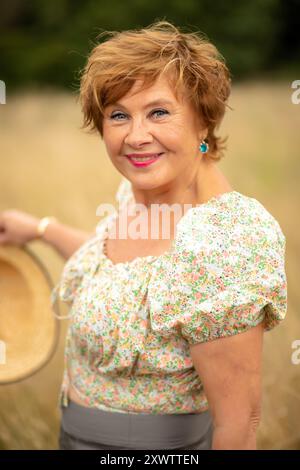 Une femme joyeuse se tient debout dans un pré doré, tenant un panier, vêtue d'un haut floral, avec la lumière du soleil illuminant son expression joyeuse. Banque D'Images