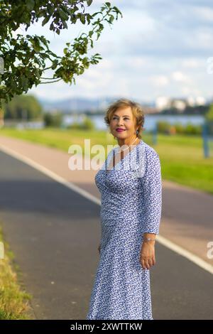 Une femme dans une robe florale bleue se tient gracieusement au bord de la rivière, profitant du plein air par temps clair. Banque D'Images