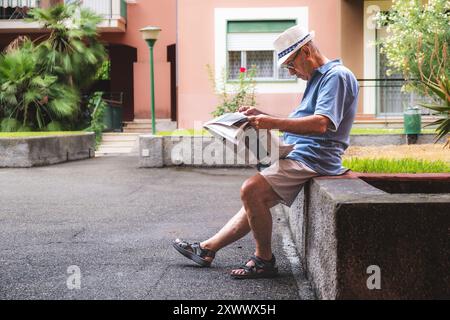 Un homme plus âgé est assis sur un mur avec un journal sur ses genoux - homme âgé portant un chapeau et une chemise bleue. La scène est paisible et calme, avec le Banque D'Images