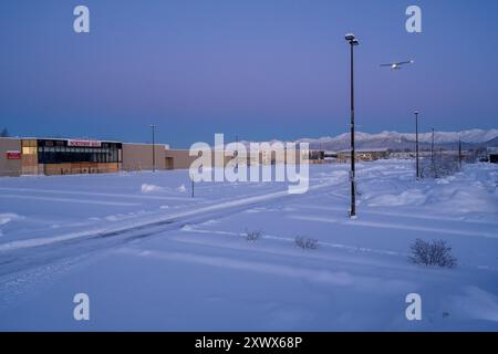 Un avion glisse gracieusement au-dessus du paysage enneigé près du centre commercial Northway Mall et de l'aéroport Merrill Field à Anchorage, en Alaska. La scène hivernale sereine capture le calme et l'étendue d'une journée enneigée. Banque D'Images