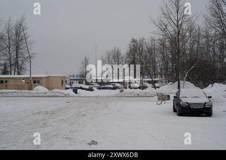 Parc de caravanes enneigé à Anchorage, Alaska, mettant en valeur la rudesse et la solitude de l'hiver. Comprend des voitures stationnées, des arbres arides et un chariot abandonné, symbolisant l'isolement et la résilience. Banque D'Images