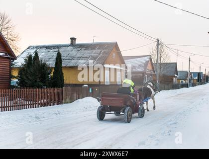 Une scène hivernale sereine représentant un traîneau tiré par des chevaux traversant une route de village enneigée près du parc national de Białowieża. L'image capture un style de vie rural tranquille, évoquant un sentiment de nostalgie et de simplicité. Banque D'Images