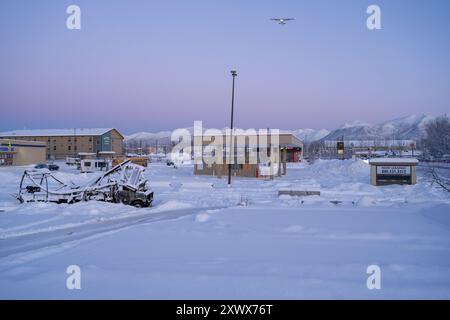 Un paysage enneigé serein près de l'aéroport de Merrill Field à Anchorage, Alaska, capturé à l'aube. Un petit avion est vu dans le ciel, avec une station-service, des bâtiments et des montagnes en arrière-plan, représentant l'isolement et la tranquillité. Banque D'Images