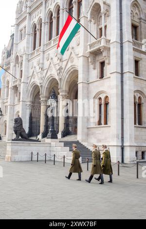 Trois gardes en uniforme défilent avec diligence devant le grand bâtiment du Parlement hongrois, mettant en valeur le mélange d'histoire et d'architecture. La majestueuse structure témoigne du riche patrimoine culturel de Budapest, avec un drapeau hongrois agitant ajoutant une fierté nationale à la scène. Banque D'Images