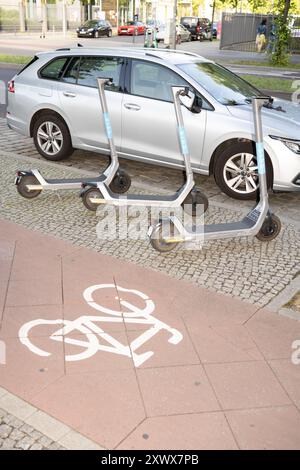 Trois scooters électriques sont garés sur une piste cyclable, bloquant la zone cyclable désignée, près d'une voiture garée. La scène se déroule dans une rue de la ville de Berlin, mettant en évidence les défis du transport urbain. Banque D'Images