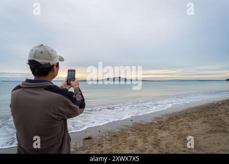 Homme prenant des photos à l'aide d'un smartphone. Île de Rangitoto au lever du soleil. Milford Beach. Auckland. Banque D'Images