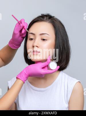 Fille adolescente Brunette ayant un tatouage de maquillage permanent sur ses sourcils. Le maquilleur fait des marques avec de la pâte blanche pour le tatouage des sourcils. Professio Banque D'Images