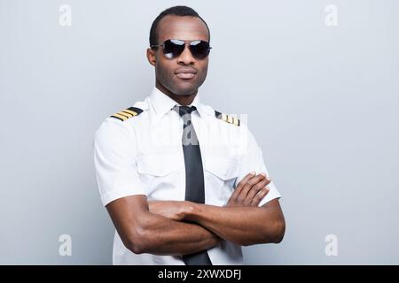 Pilote confiant et expérimenté. Pilote africain confiant en uniforme gardant les bras croisés tout en se tenant debout sur fond gris Banque D'Images