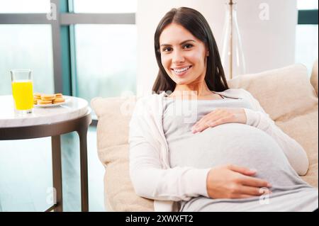 Bientôt disponible... Belle jeune femme enceinte touchant son abdomen et souriant tout en étant allongée sur le canapé à la maison Banque D'Images