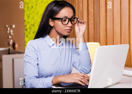 Femme d'affaires confiante. Attirante jeune femme africaine travaillant sur ordinateur portable et ajustant ses lunettes tout en étant assise à son lieu de travail Banque D'Images