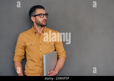 Perdu dans les pensées d'affaires. Homme mûr réfléchi transportant un ordinateur portable et regardant loin tout en se tenant debout sur fond gris Banque D'Images