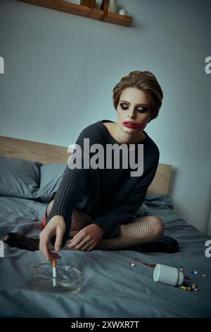 Une jeune femme est assise sur un lit, les yeux baissés, entourée de pilules éparpillées et d'un cendrier. Banque D'Images