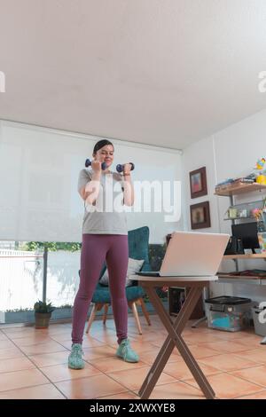 Belle jeune femme latina qui s'exerce dans son salon devant son ordinateur portable suite à un tutoriel vidéo pendant la journée Banque D'Images