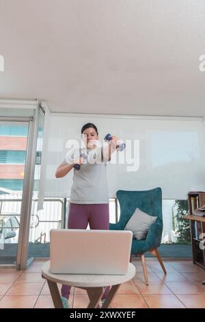 Belle jeune femme latina qui s'exerce dans son salon devant son ordinateur portable suite à un tutoriel vidéo pendant la journée Banque D'Images