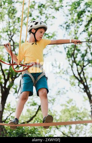 Un garçon dans un parc de cordes dans la nature apprend à grimper. Un enfant dans un casque marche le long d'un téléphérique surmontant les obstacles. Banque D'Images