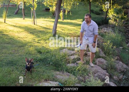 Souriant homme d'âge moyen marche et joue avec un petit chien dans un parc public Banque D'Images