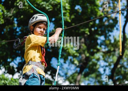 Un garçon dans un parc de cordes dans la nature apprend à grimper. Un enfant dans un casque marche le long d'un téléphérique surmontant les obstacles. Banque D'Images