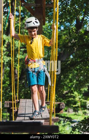 Un garçon dans un parc de cordes dans la nature apprend à grimper. Un enfant dans un casque marche le long d'un téléphérique surmontant les obstacles. Banque D'Images