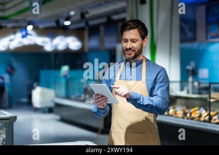 Employé de supermarché dans le tablier en utilisant la tablette numérique pour la gestion des stocks. L'image représente la technologie dans le commerce de détail, l'efficacité et l'environnement moderne des épiceries. Produits frais et cadre vibrant Banque D'Images