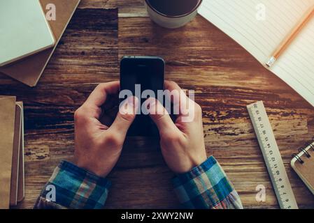 Envoi d'un SMS professionnel. Vue de dessus en gros plan de mains masculines tenant le téléphone portable sur un bureau en bois avec différentes choses chancellerie posées sur elle Banque D'Images