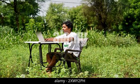 Jeune garçon indien utilisant un ordinateur portable à l'extérieur. Homme assis avec table de bureau au jardin et faisant du travail à la maison. Banque D'Images